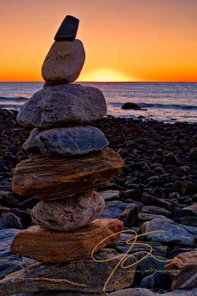 Neatly stacked, one atop the other, a rock cairn sitting among the boulders on the rocky coast at Rye Harbor. The sun is just about to peak above the horizon giving the cloudless sky a nice ogange glow