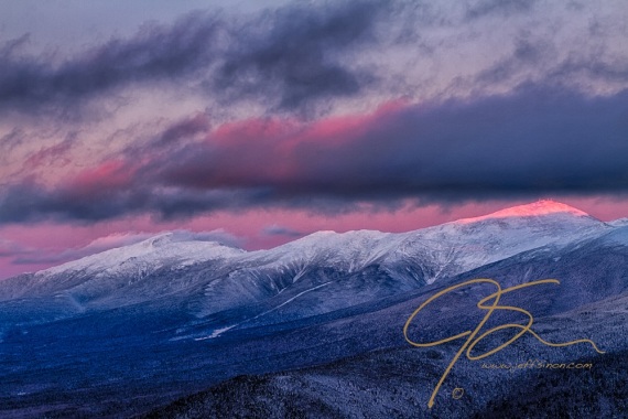 Alpenglow reaches the summit of Mount Washington, piercing through a gap in the clouds. The snowy peaks of the rest of the Presidential Range trailing off into the distance towards image left.